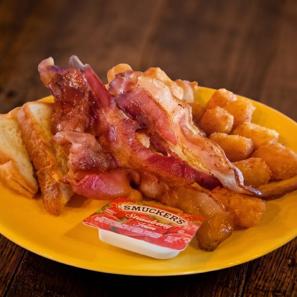 A breakfast plate with crispy bacon, golden tater tots, toast, and strawberry jam.