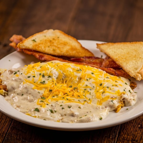 Breakfast plate with eggs and gravy topped with cheese, bacon, and toast on a wooden table.