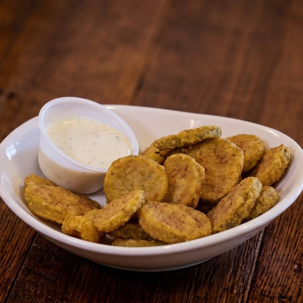 White bowl of golden fried pickle slices with a side of creamy dipping sauce on a wooden table.