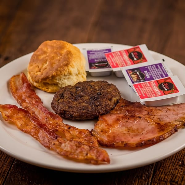 A breakfast plate with a biscuit, sausage, bacon, ham, and jam packets on a wooden table.