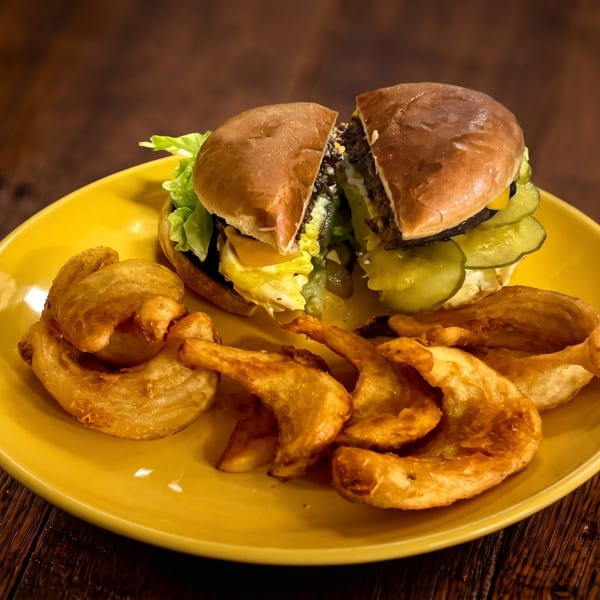 Half-cut burger with lettuce, pickles, and cheese, served with curly fries and potato wedges on a yellow plate.