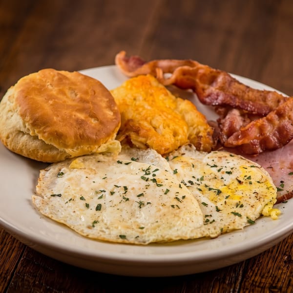 Hearty breakfast plate with fried eggs, biscuit, hash browns, bacon, and ham on a wooden table.