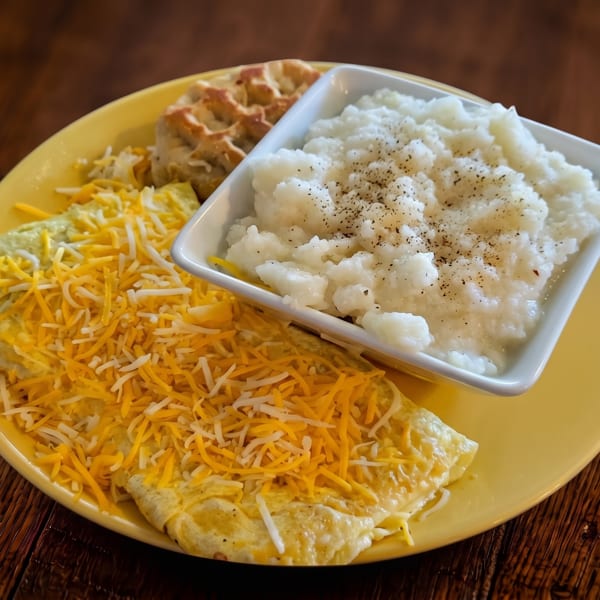 A yellow plate holds a cheesy omelet, peppered grits, and a waffle on a dark wooden table.